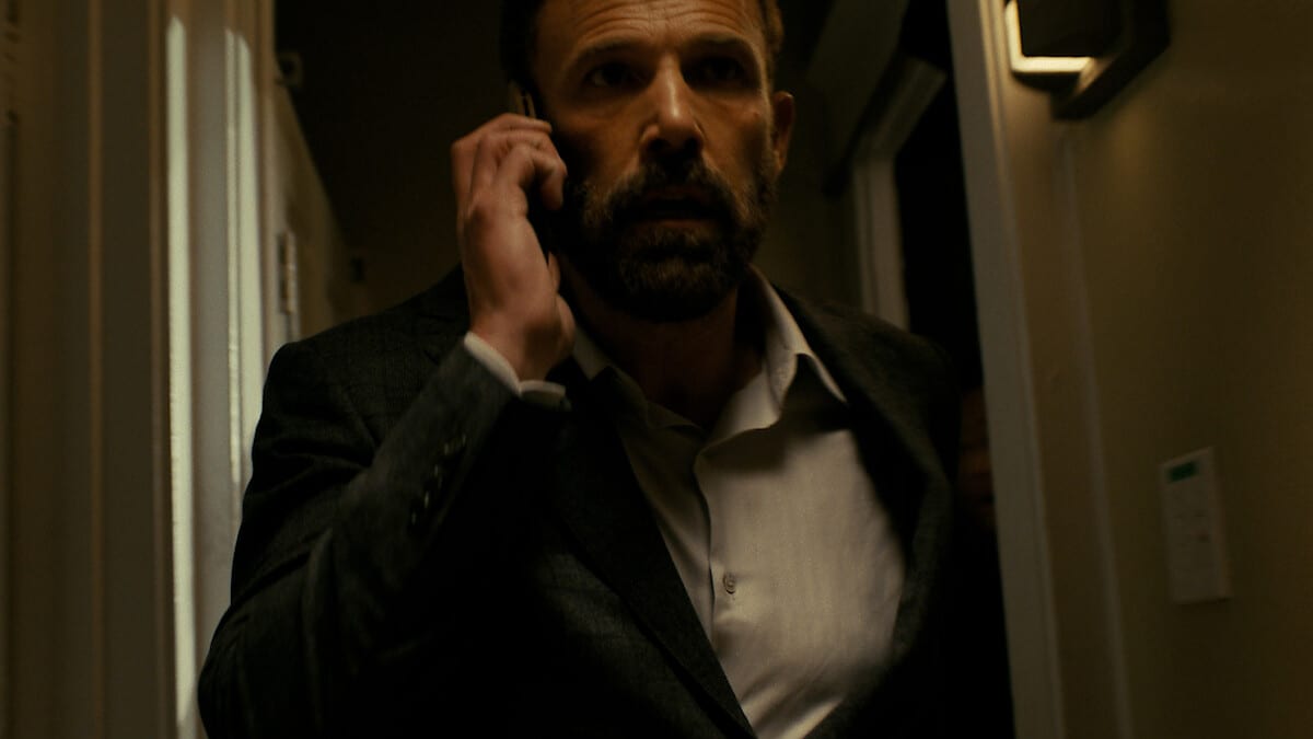A man in a dark suit talks on the phone as he walks down a dimly lit hallway with warmly lit walls.