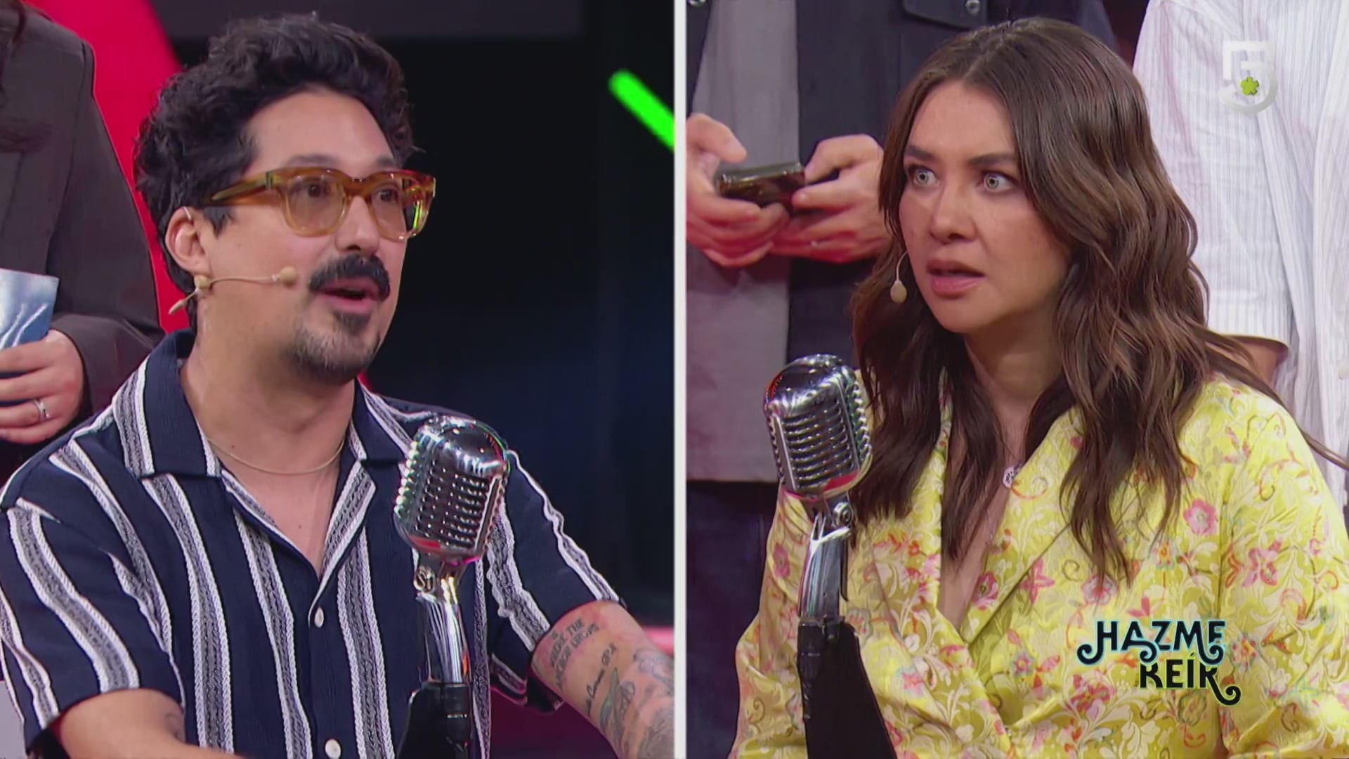 Pantalla dividida en dos con un hombre de lentes y bigote que habla a la izquierda y una mujer de cabello castaño que mira con sorpresa a la derecha, ante micrófonos de época.