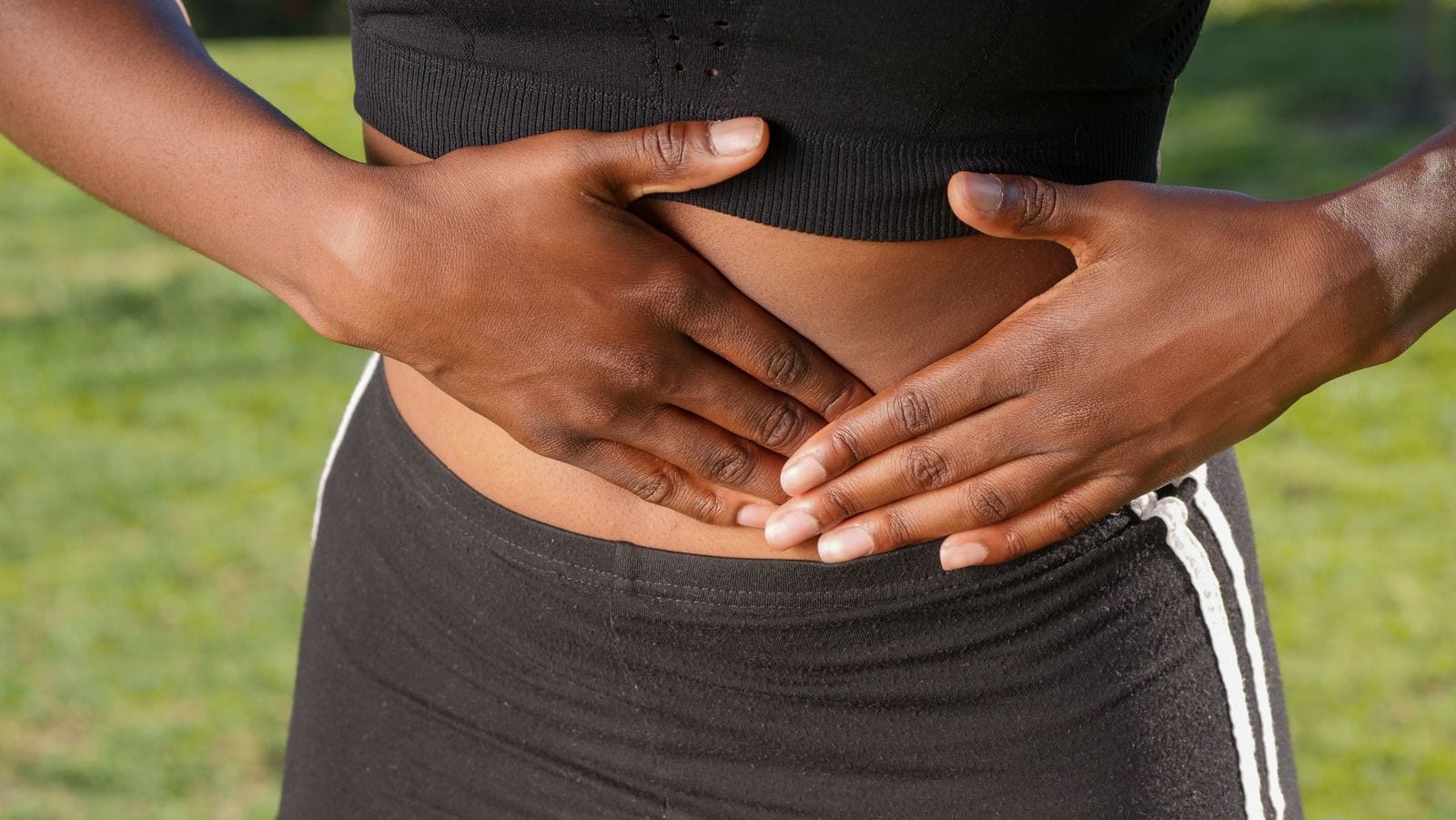 Las dos manos de una persona sobre el abdomen, con ropa deportiva negra y pasto al fondo.