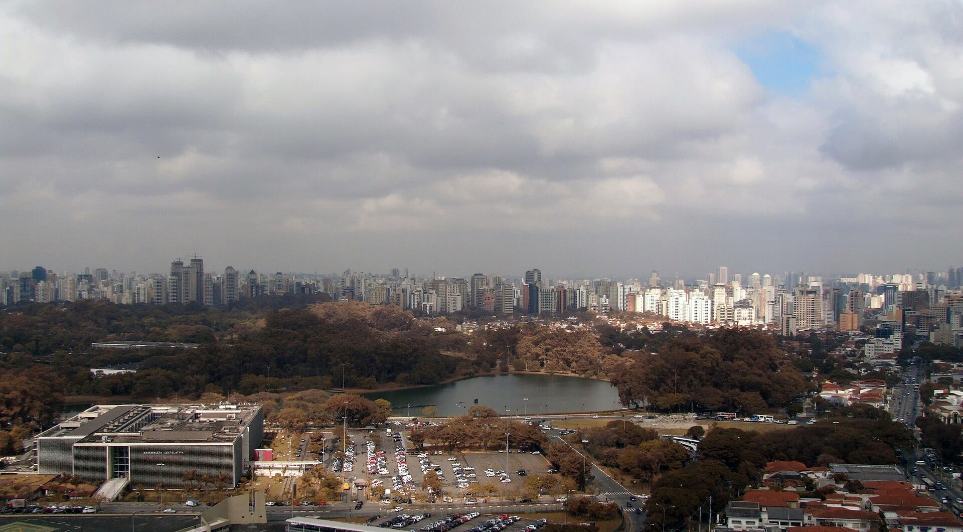Vista aérea del edificio de la Asamblea Legislativa de São Paulo junto a un estacionamiento y un lago, con los rascacielos de la ciudad al fondo.