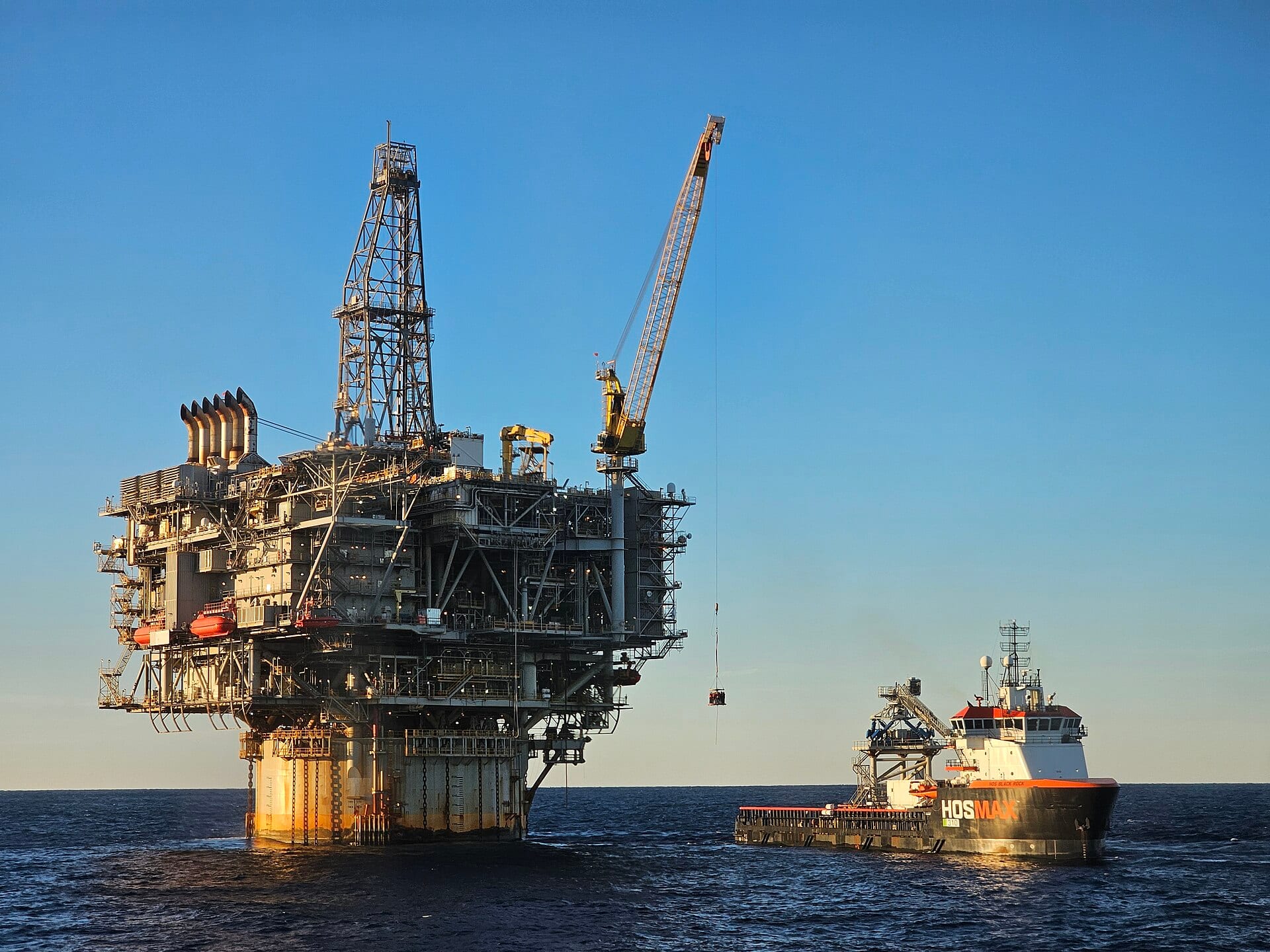 Una plataforma petrolera costa afuera con grúa y torre de perforación, junto a un buque de suministro, bajo un cielo despejado.
