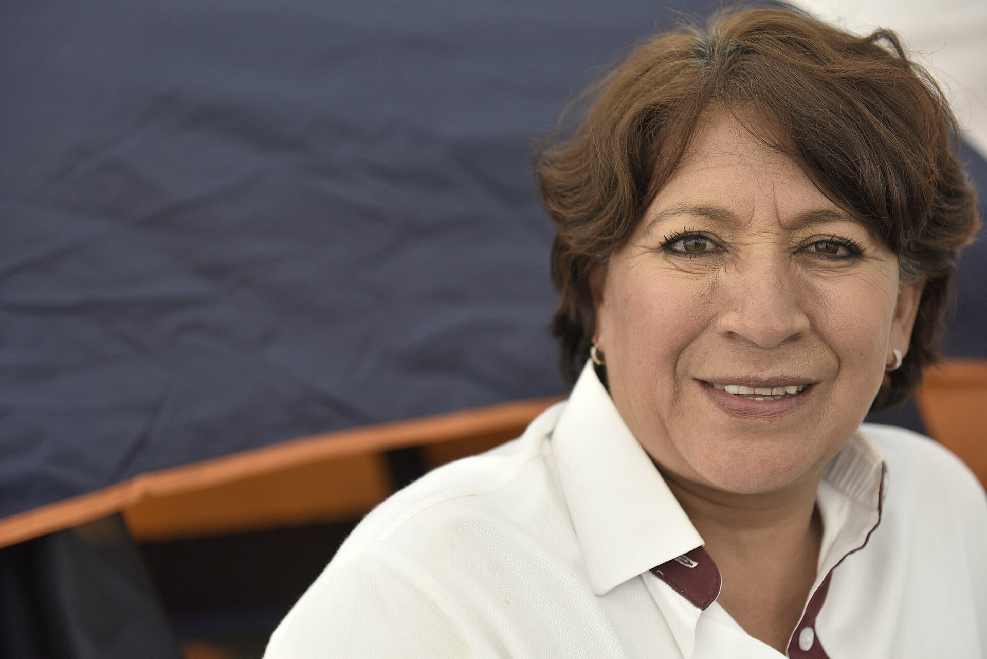 Delfina Gómez sonríe a la cámara con una camisa blanca de cuello guinda, frente a una lona azul y una banca de madera.