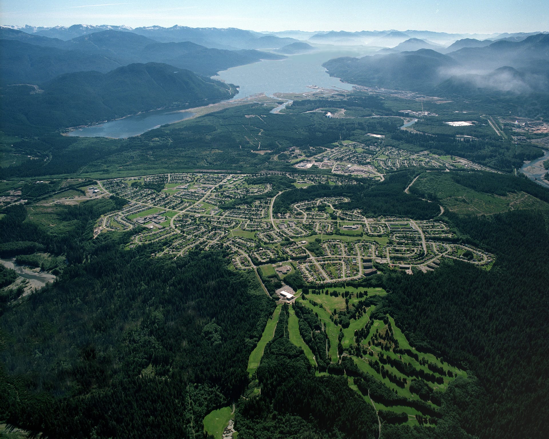 Vista aérea de un poblado de calles curvas rodeado de bosque denso, con un canal de agua y montañas nubladas al fondo.