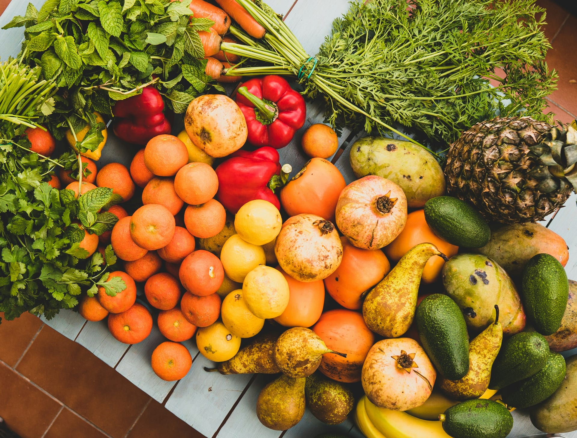 A pile of fruit and vegetables on a table seen from above, with oranges, lemons, pomegranates, red peppers, avocados, a pineapple and bunches of herbs.