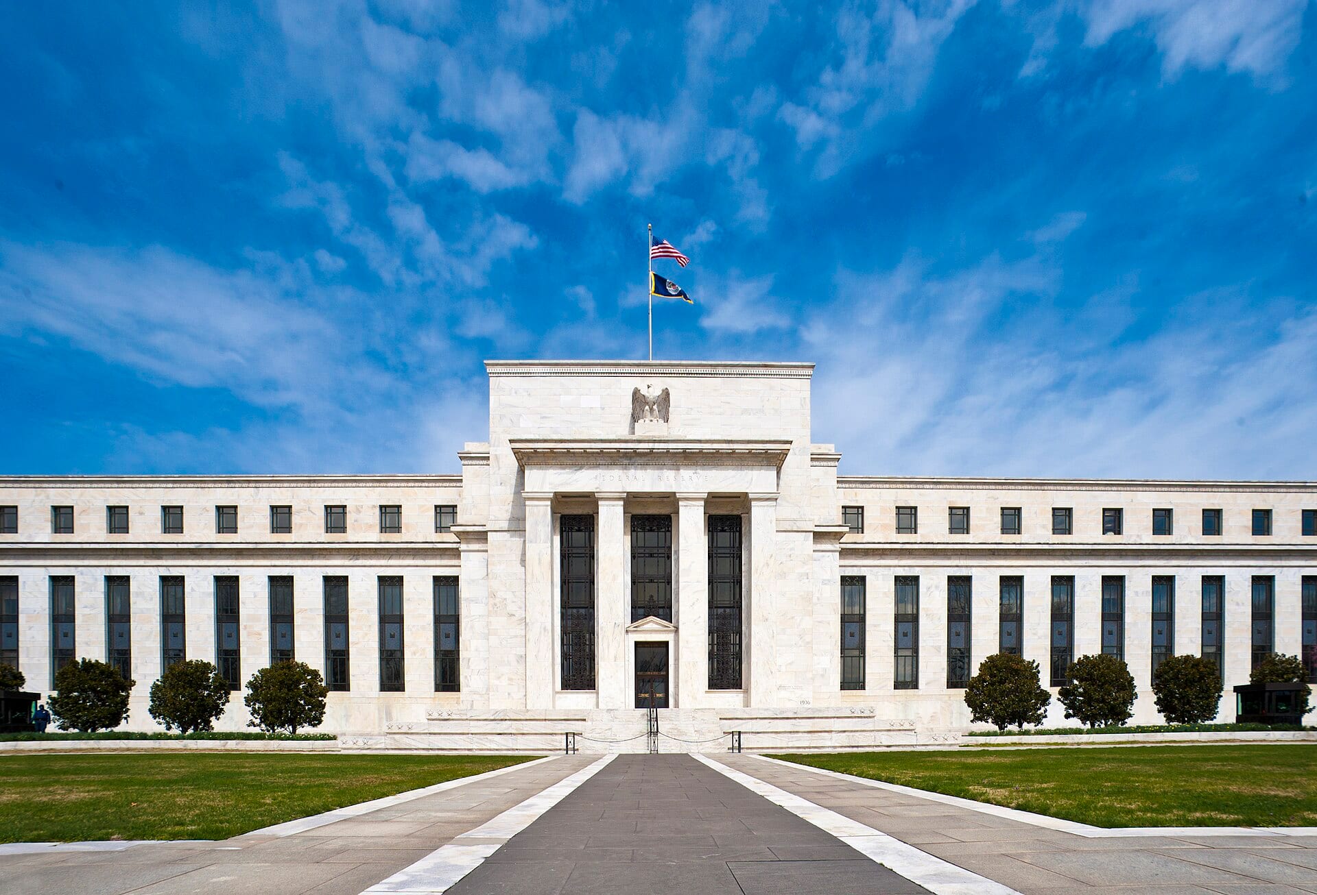 The white marble facade of the Federal Reserve's Eccles Building in Washington, with a U.S. flag above the portico under a blue sky with clouds.