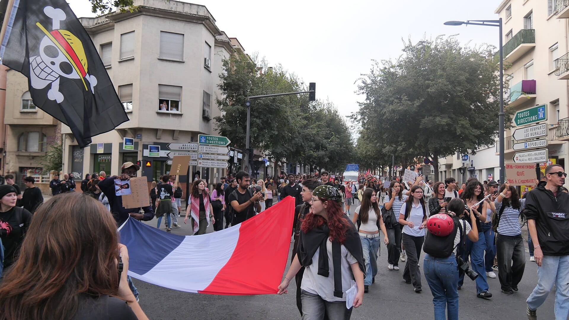 Young people march down a street holding a stretched French tricolor flag and a black pirate-skull flag, among cardboard signs.