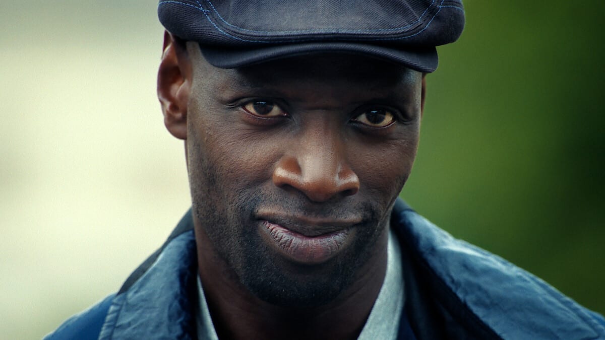 Primer plano de Omar Sy como Assane Diop, con gorra oscura y chamarra azul, mirando a la cámara con una leve sonrisa.