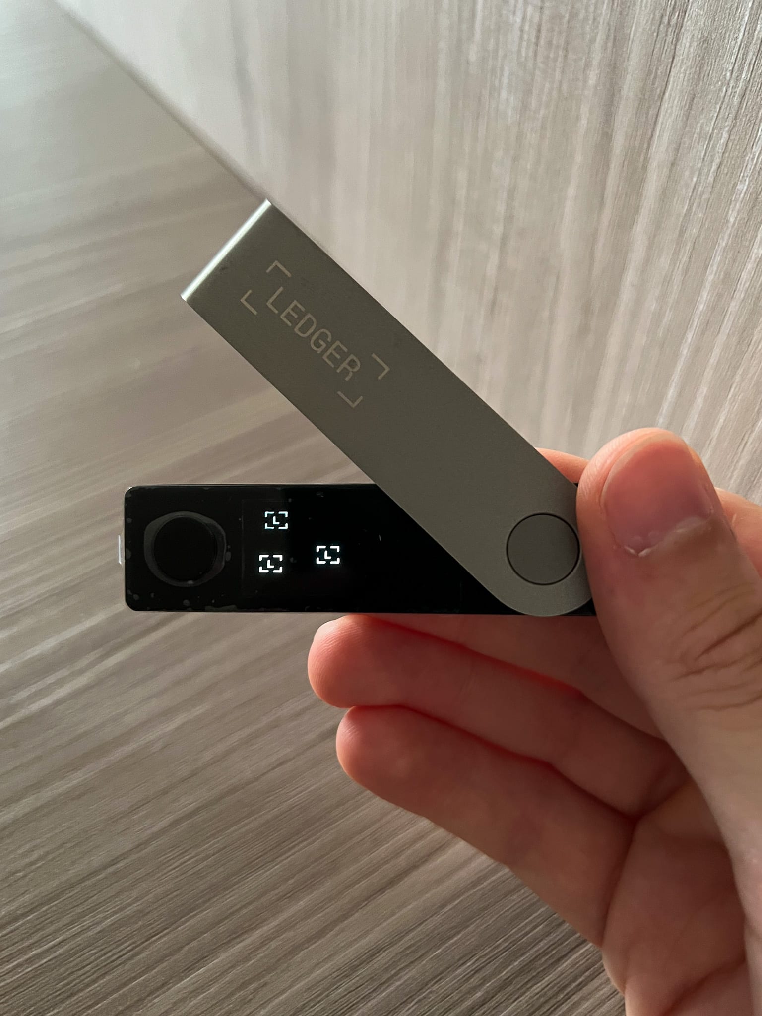 A hand holds a Ledger Nano X crypto wallet with its metal cover raised and the screen lit.