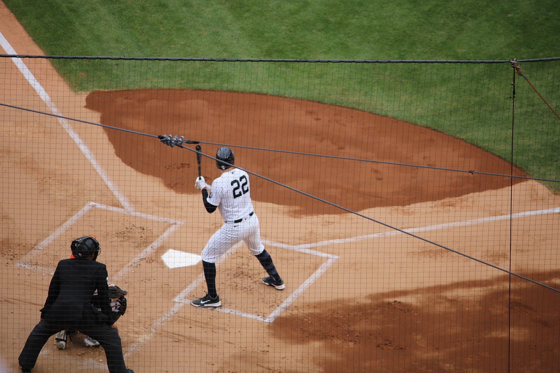Un bateador con el número 22 de los Yankees, visto de espaldas, espera el lanzamiento junto al home mientras el umpire se agacha detrás de una red.