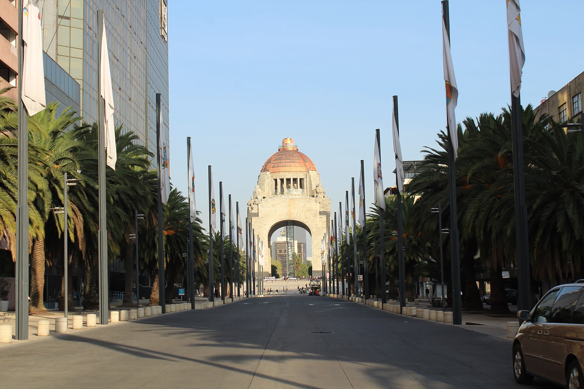 El Monumento a la Revolución, con su cúpula de cobre, visto al fondo de una avenida con palmeras y astas con banderas.