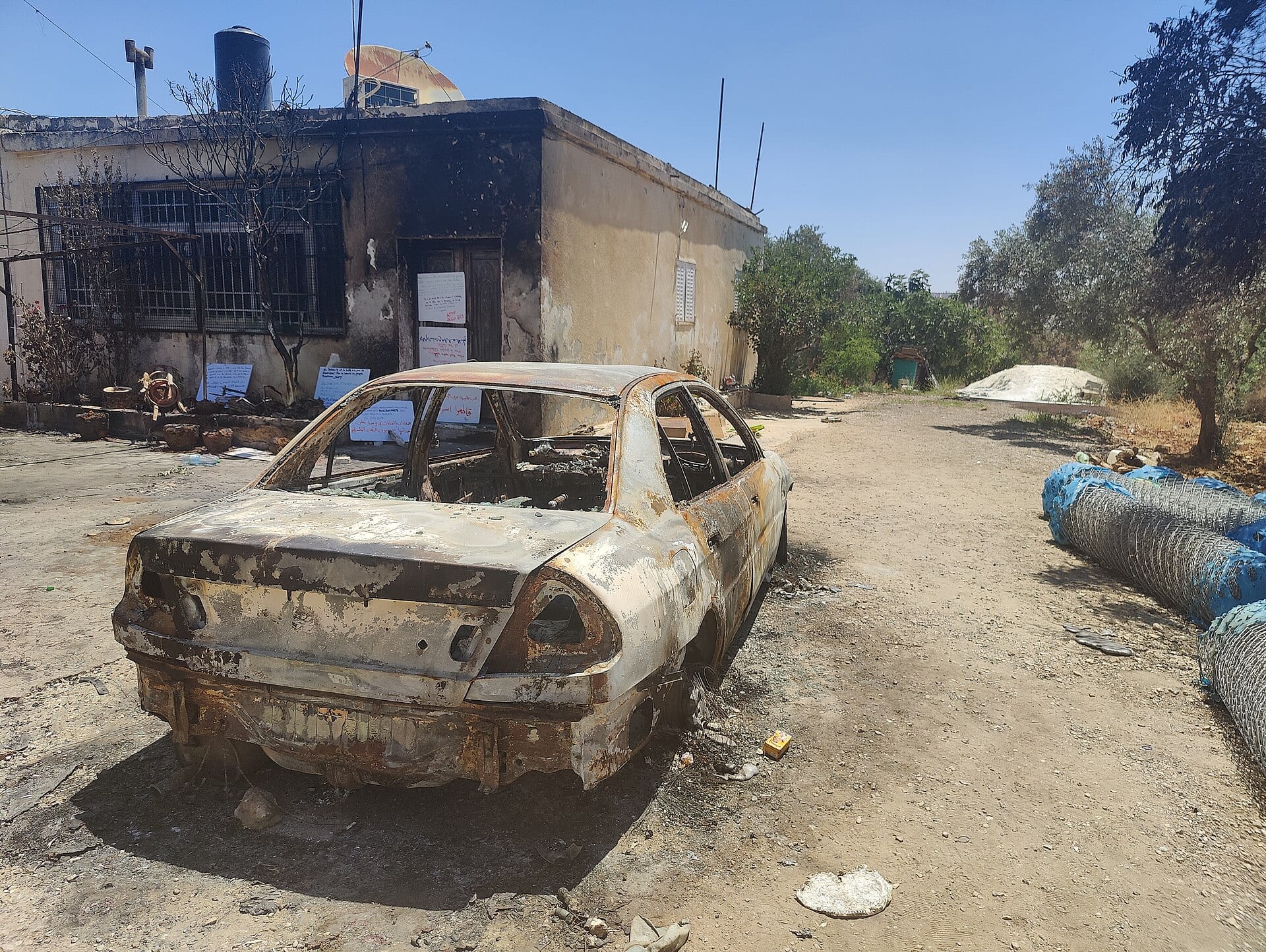 A burned-out car in front of a house with blackened walls, next to an olive tree, on a dirt road.