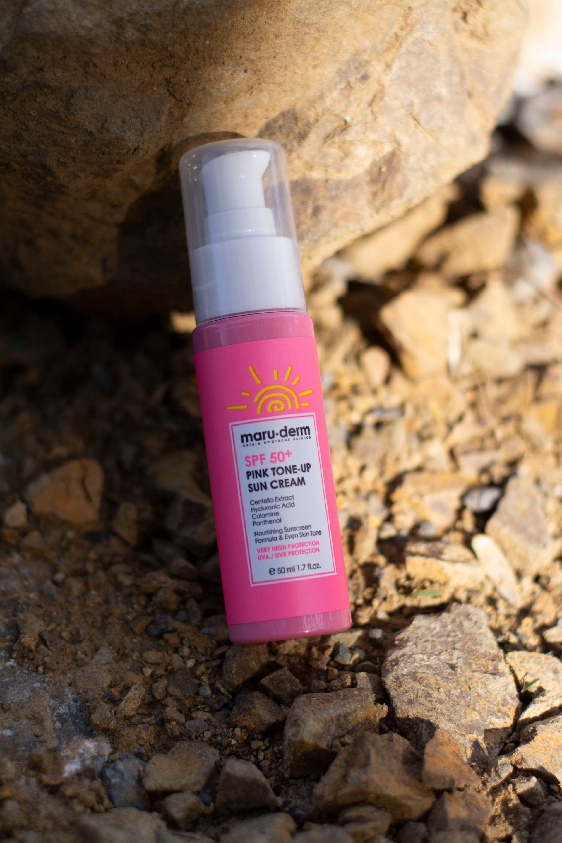 Un frasco rosa de crema con protector solar SPF 50+ apoyado sobre piedras, al aire libre.