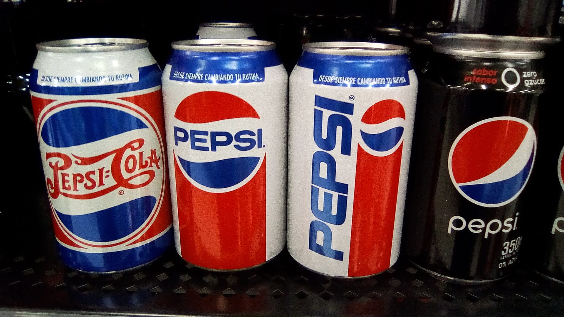 Cuatro latas de Pepsi en un refrigerador, tres con diseños de logotipo de distintas épocas y una negra de Pepsi Zero Azúcar.