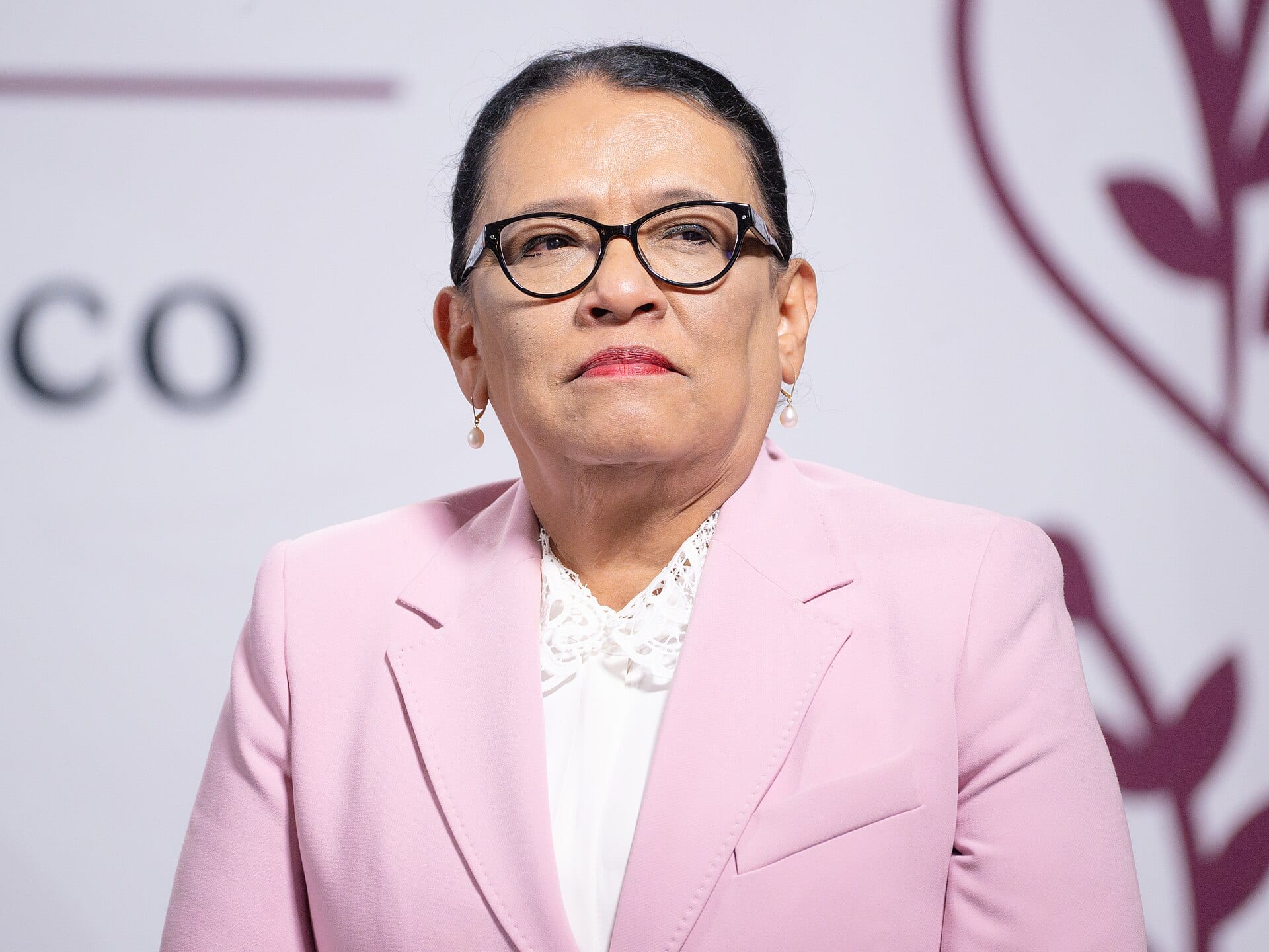 Rosa Icela Rodríguez, de saco rosa y lentes oscuros, frente a un fondo claro con motivos vegetales.