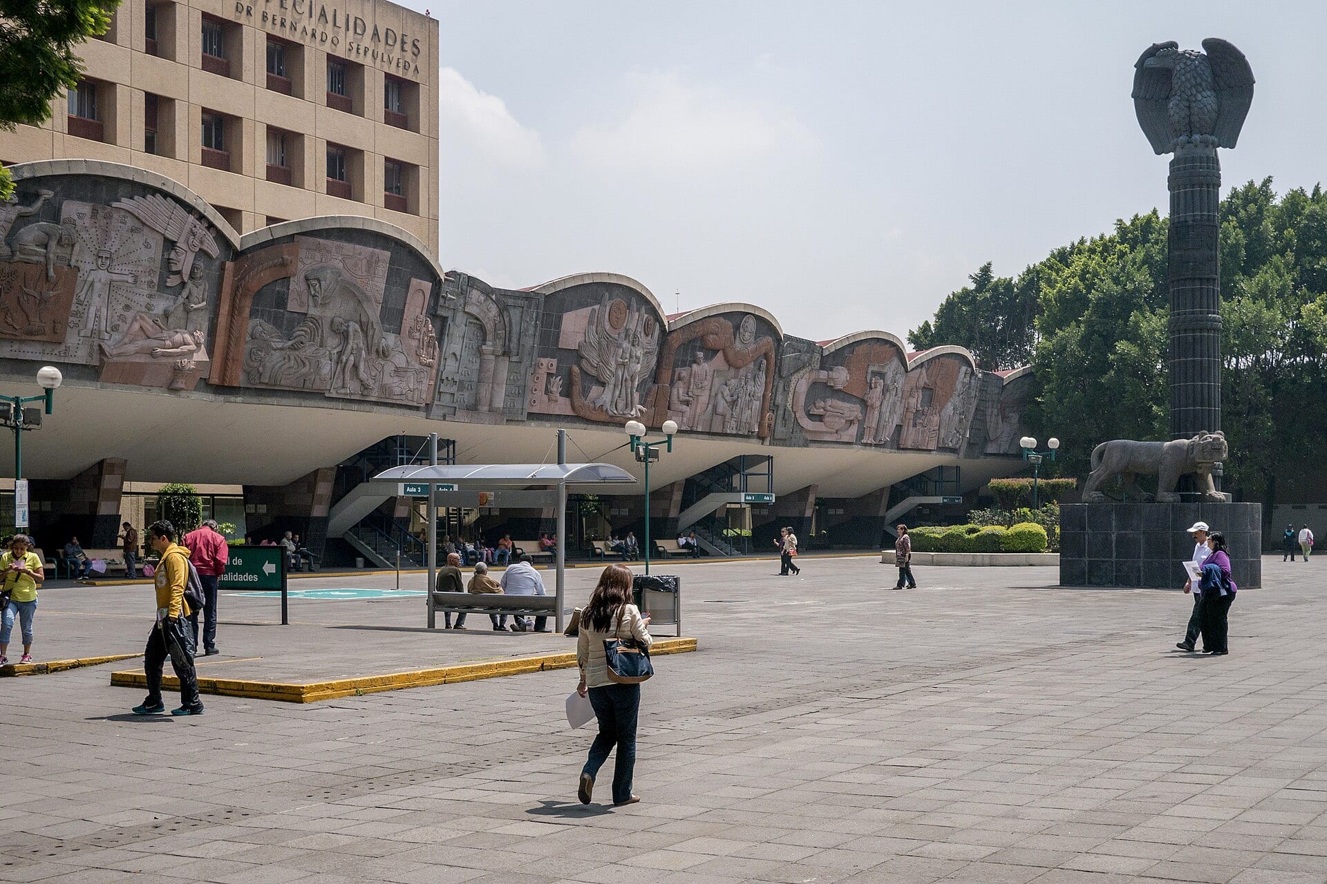 Explanada del Centro Médico Nacional Siglo XXI, con el friso escultórico del hospital de especialidades, una columna con un águila y personas caminando.