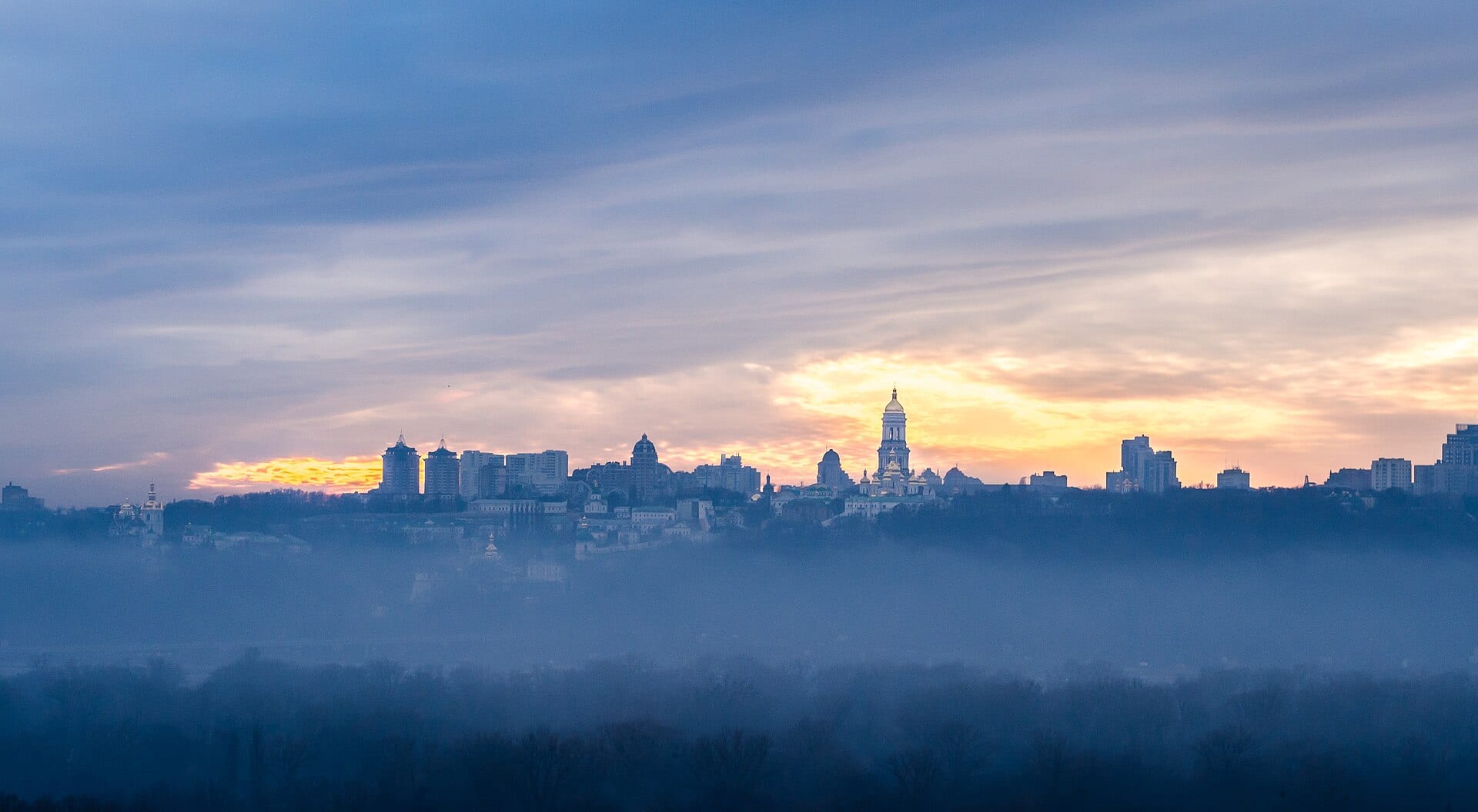 Siluetas de edificios y cúpulas de Kiev sobre una capa de niebla, con nubes anaranjadas en el horizonte al atardecer.