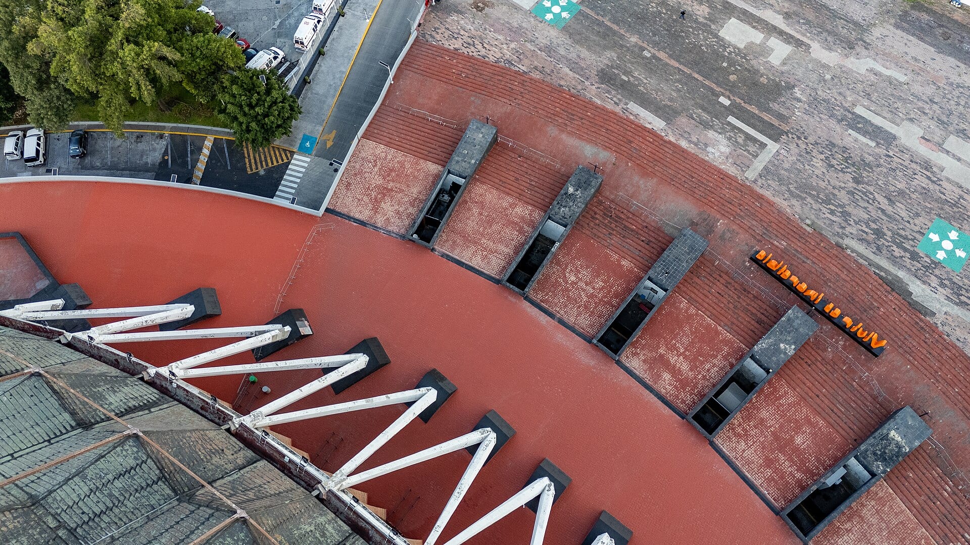Vista aérea desde arriba de la cubierta circular del Palacio de los Deportes, de color rojizo, con vigas blancas y un estacionamiento al fondo.