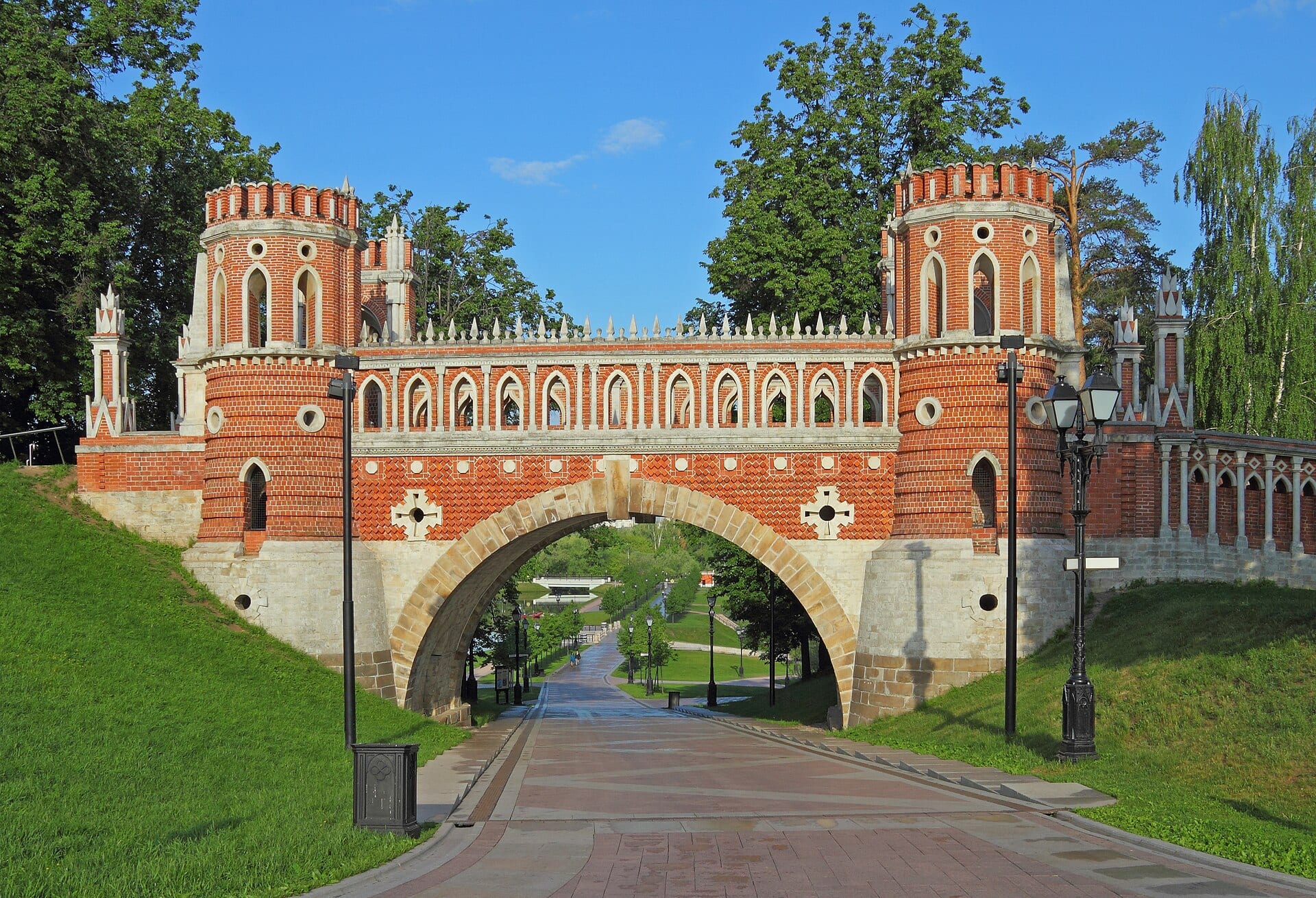 A red-brick bridge with crenellated towers and a stone arch over a path in Tsaritsyno park, under a blue sky.