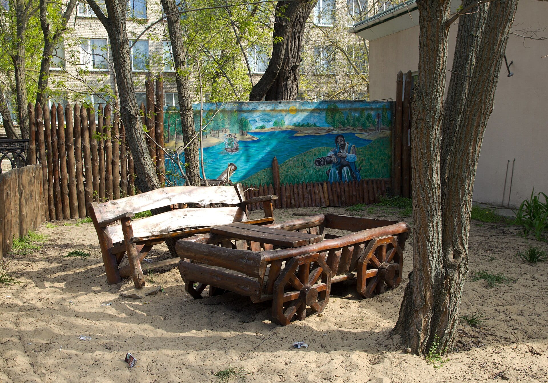 A wooden bench and table on sand under trees, in front of a mural of a river with a Cossack seated beside a cannon.