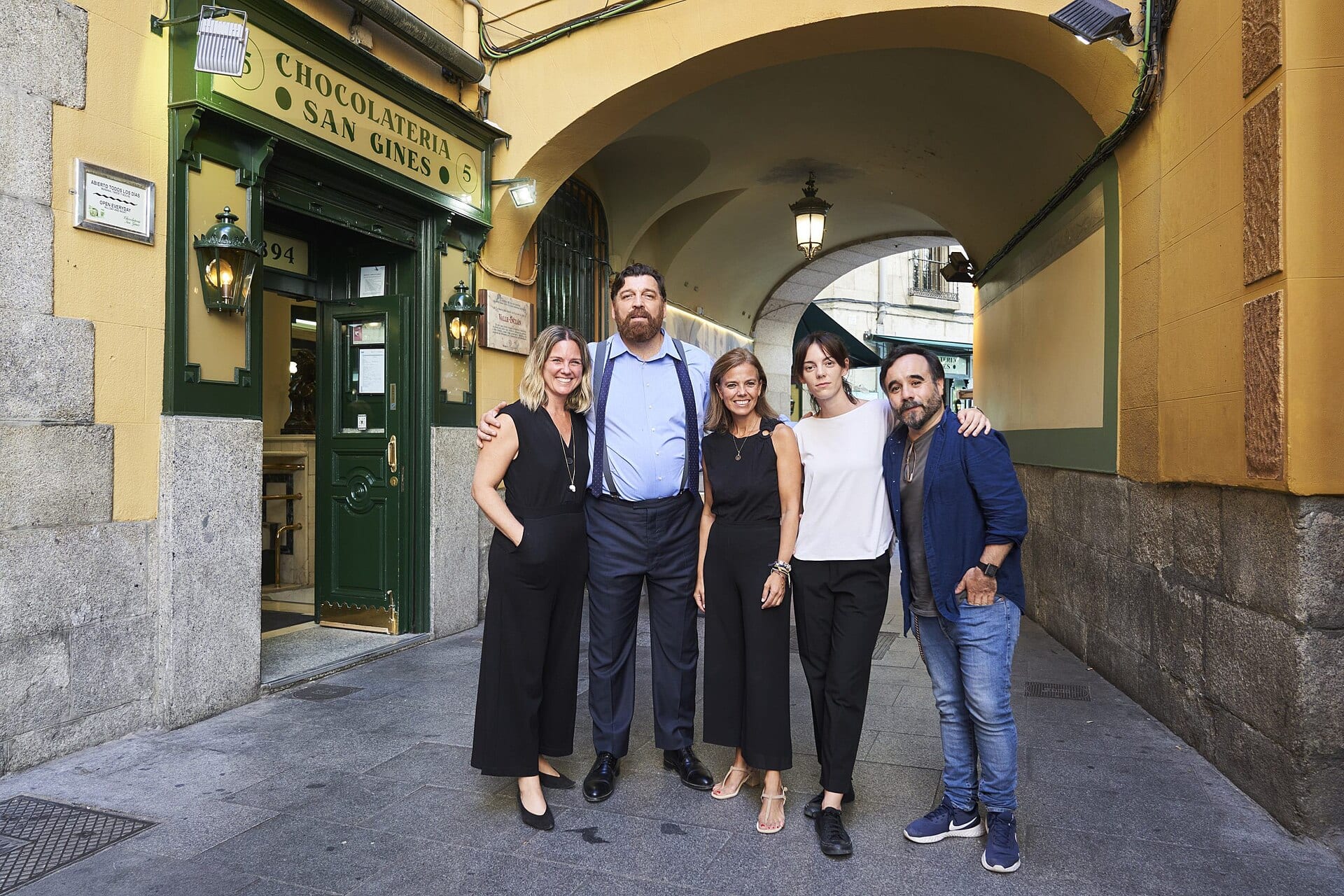 Cinco personas posan abrazadas bajo un arco de un callejón de Madrid, junto a la puerta verde de una chocolatería, en una visita al rodaje de 2022.