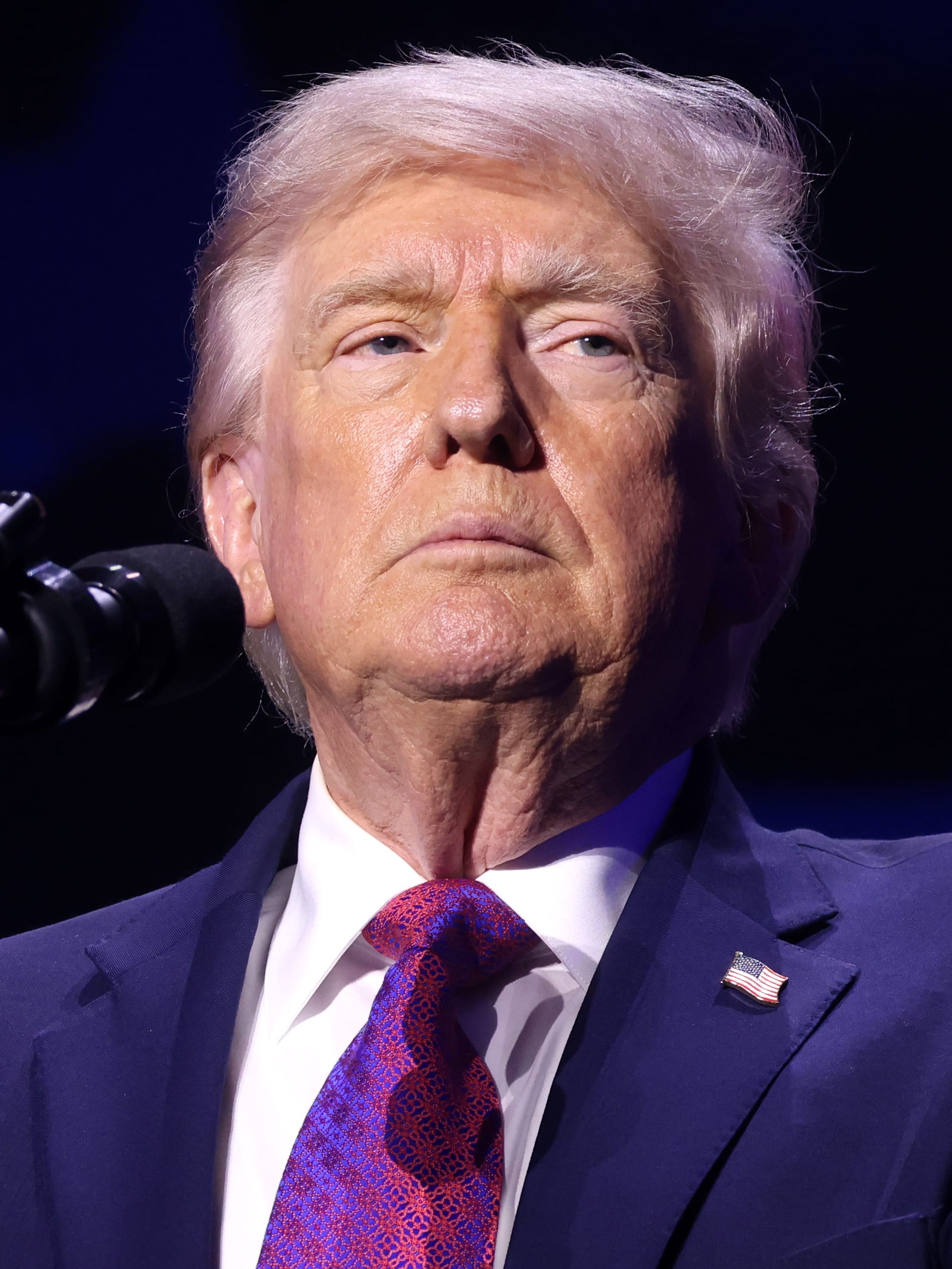 Primer plano de Donald Trump, de traje azul y corbata morada, frente a un micrófono sobre un fondo oscuro.