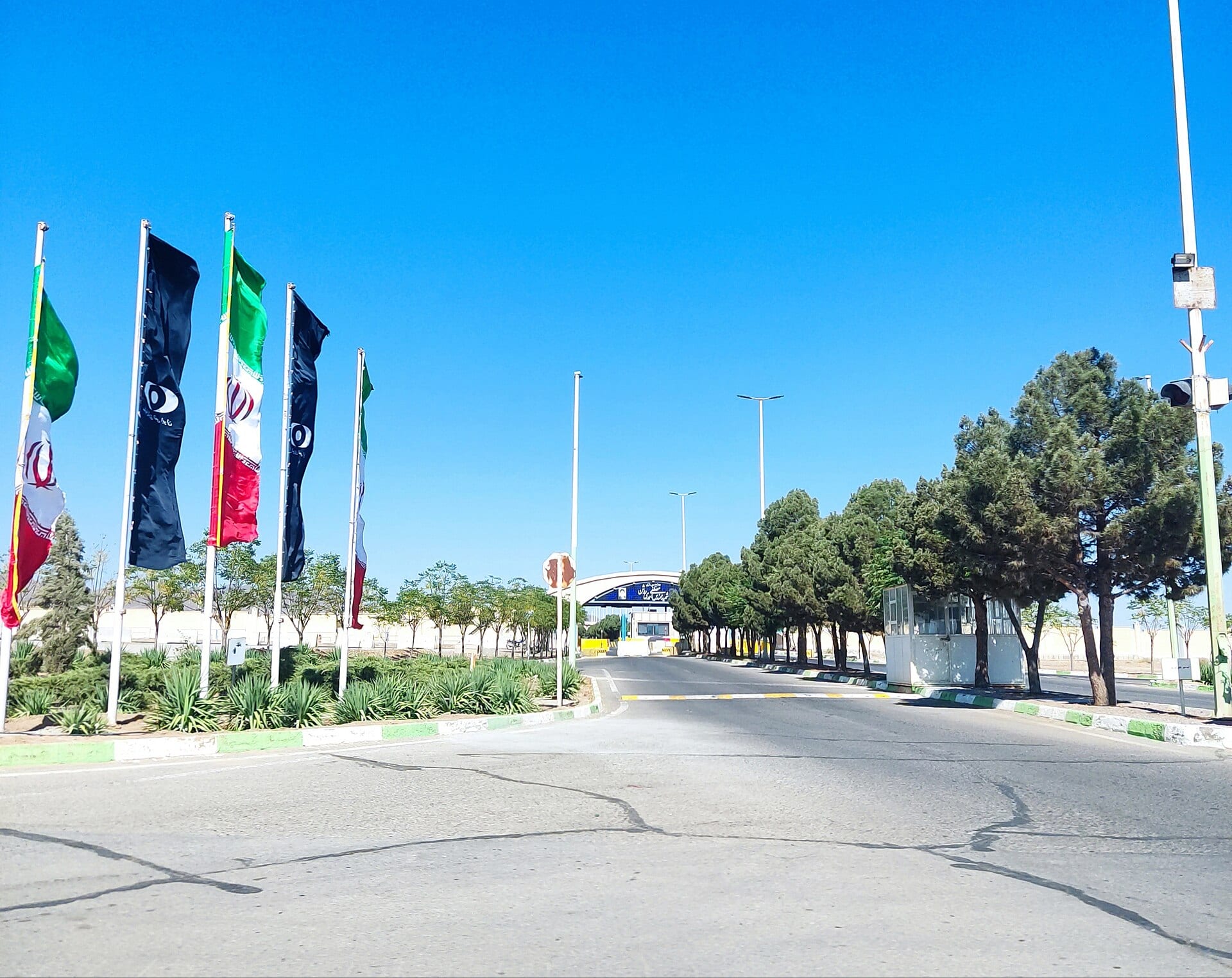 Five tall flags, three bearing an atomic emblem, beside an empty road leading to a security gate under a clear sky.