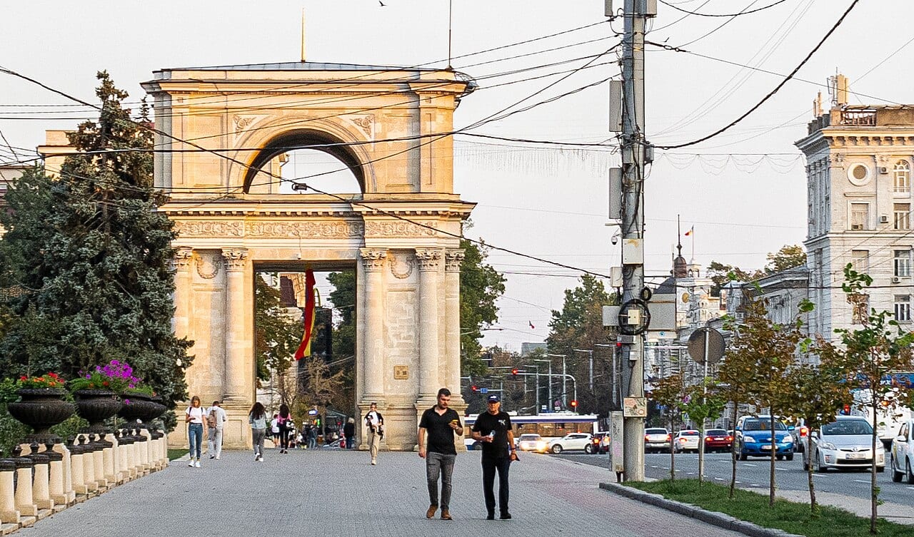 Un arco triunfal de piedra al final de una avenida de Chisináu, con peatones y autos en la calle.