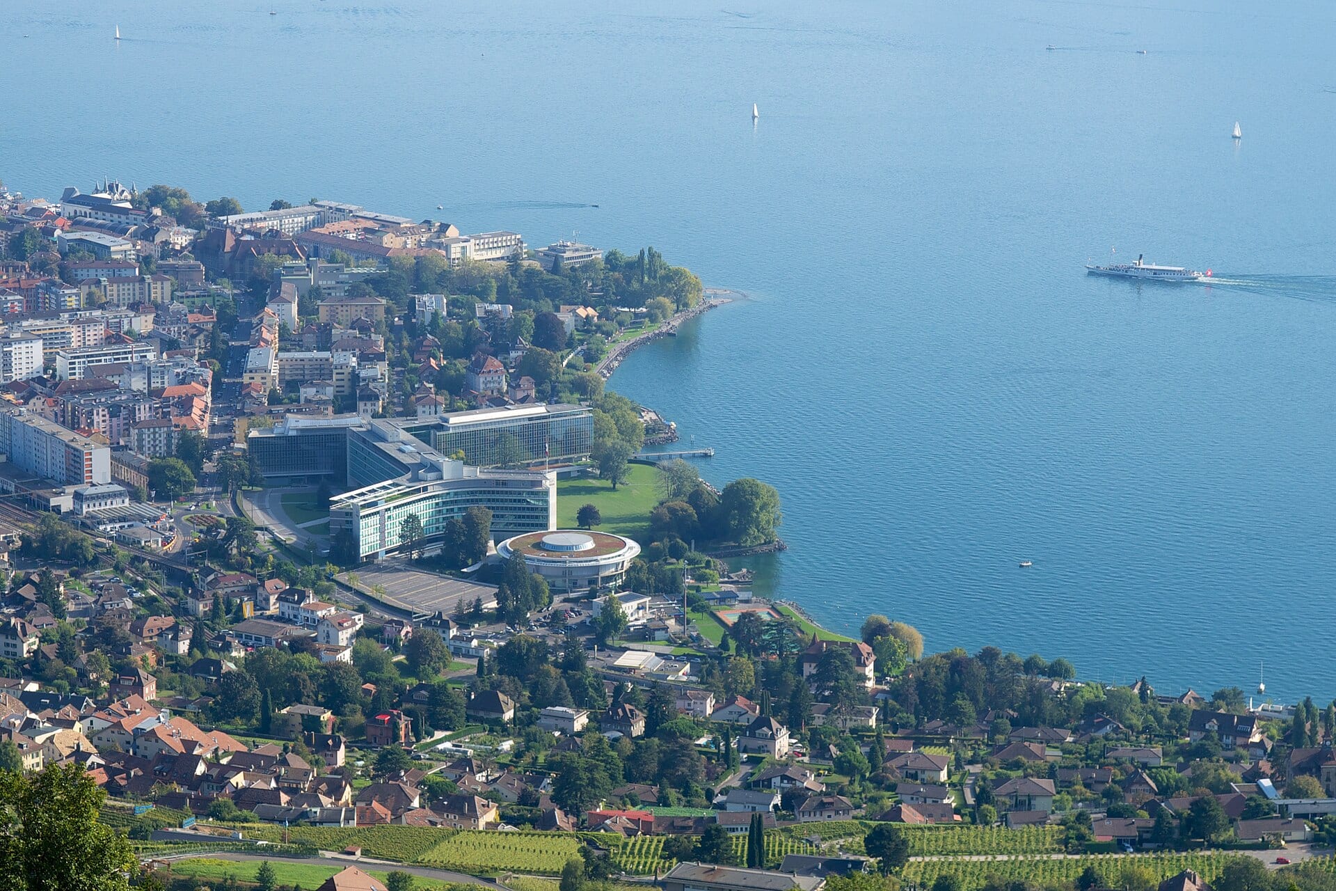 Vista aérea de la sede de Nestlé en Vevey, Suiza, con edificios de vidrio junto al lago y un barco de vapor en el agua.